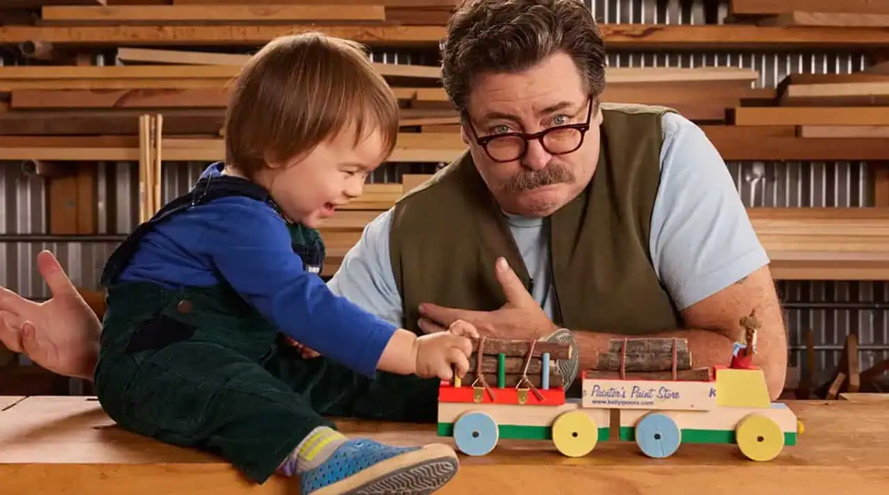Nick Offerman posing with child and wooden toy from his book "Little Woodchucks: Offerman Woodshop's Guide to Tools and Tomfoolery".